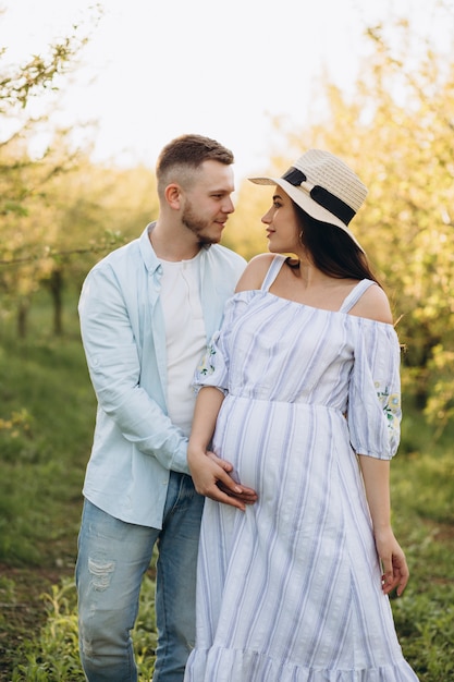 À la mode et élégante femme enceinte heureuse et son mari habillé d'un ton pastel blanc et bleu dans le jardin au coucher du soleil