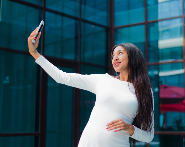 Mode femme afro dans une luxueuse robe blanche faisant selfie avec smartphone contre un bâtiment aux fenêtres bleues