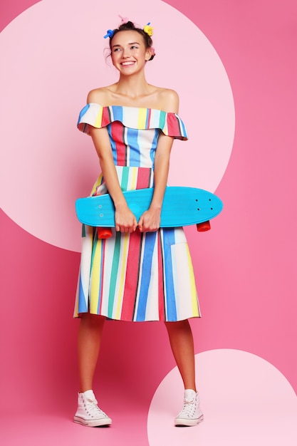 Mode femme cool posant avec une planche à roulettes, une robe multicolore et des baskets sur le rose.