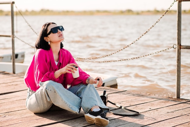 Mode femme à lunettes de soleil assis sur la jetée et fumer ecigarette en journée ensoleillée