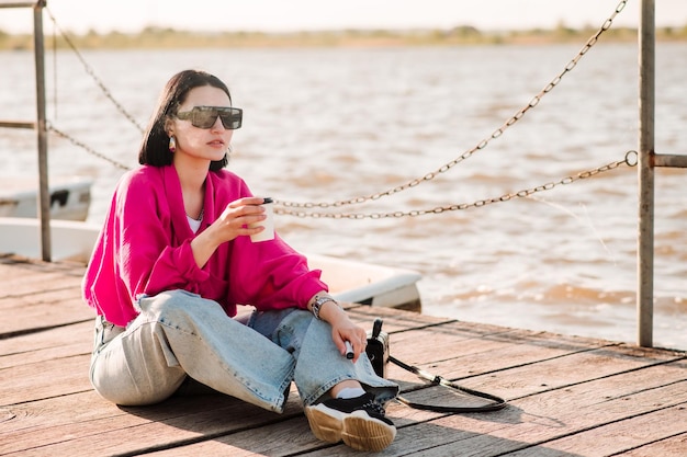 Mode femme à lunettes de soleil assis sur la jetée et fumer ecigarette en journée ensoleillée