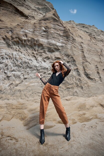 Mode fille aux cheveux roux en jeans orange posant dans la nature près de rochers de sable, pas comme tout le monde