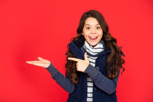 Mode d'hiver doigt pointé et présentation de l'espace de copie enfant choqué aux cheveux bouclés en gilet puffer adolescente sur fond rouge portrait d'enfant portant des vêtements chauds exprimer une émotion positive