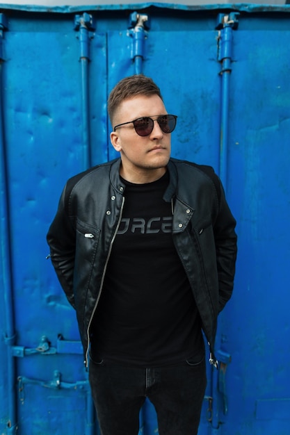 Mode jeune beau garçon avec des lunettes de soleil noires en jeans veste en cuir et t-shirt noir se tient près d'un fond bleu métal