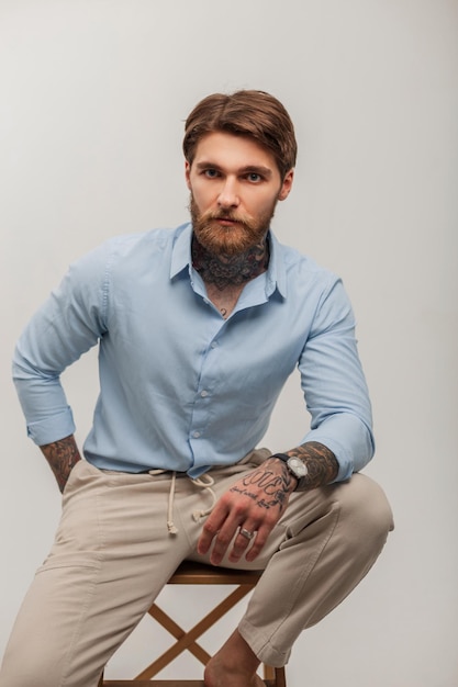À la mode jeune bel homme brutal avec barbe et coiffure avec des tatouages dans des vêtements élégants avec chemise est assis sur une chaise en bois sur fond blanc