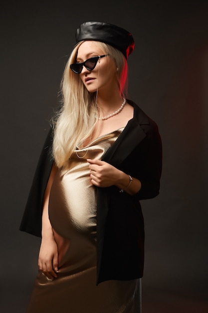 À la mode jeune femme enceinte dans une robe or, des lunettes de soleil et un chapeau à la mode sur le fond sombre. Concept de style pour la grossesse.