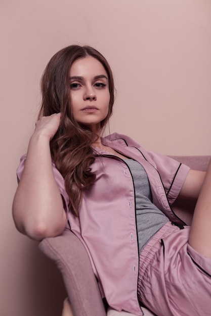 Mode jeune femme avec joli visage en vêtements de pyjama de nuit rose est assis sur une chaise près du mur rose