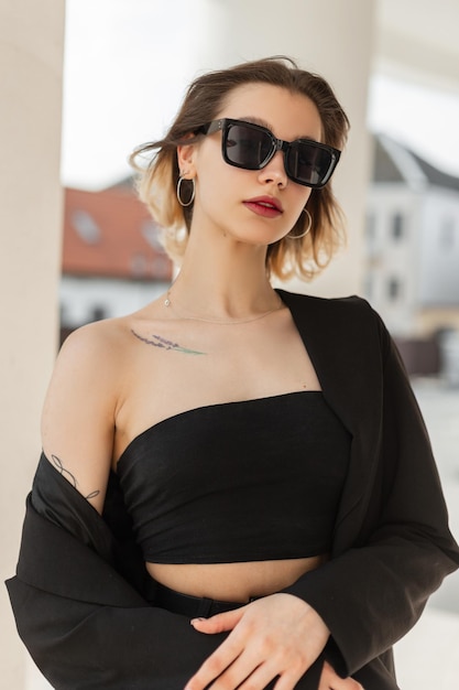 À la mode jeune mannequin élégance à la mode avec des lunettes de soleil modernes de mode dans des vêtements d'affaires noirs élégants avec haut et blazer se promène dans la ville en journée d'été ensoleillée