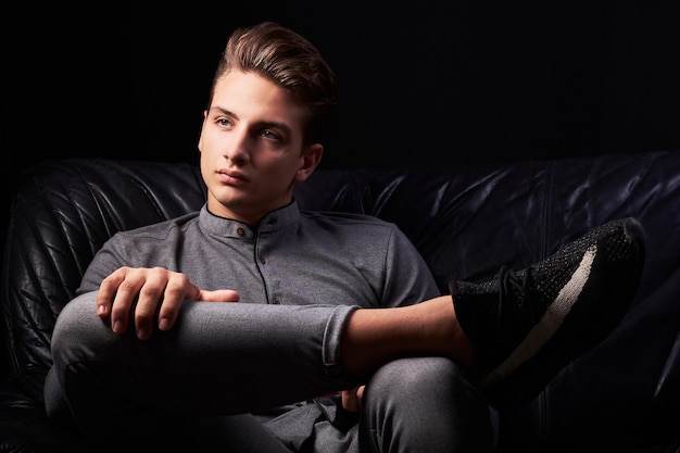 Mode portrait d'élégant jeune homme assis sur un canapé en cuir sur fond noir