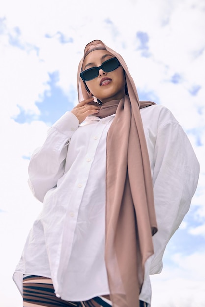 Mode portrait et une femme musulmane en plein air sur un fond de ciel bleu dans des lunettes de soleil et une écharpe pour la foi islamique de style contemporain et le hijab avec une jeune arabe à la mode à l'extérieur dans des vêtements modernes