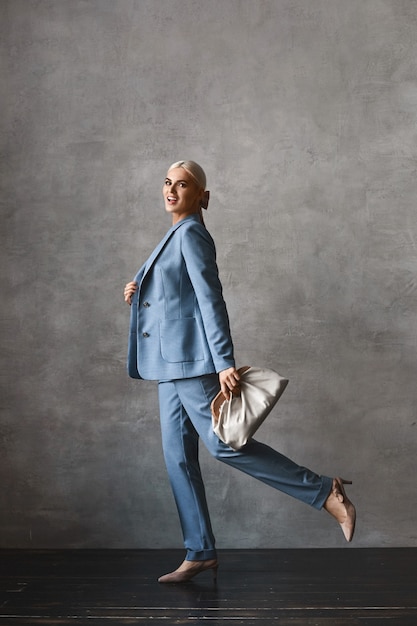 Mode portrait en pied d'une femme modèle aux cheveux blonds en costume tendance et avec un sac à main élégant à la recherche de l'appareil photo et posant à l'intérieur