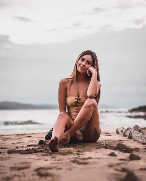 Mode posant d'une femme brune en maillot de bain et un paréo sur la plage en été assis sur le sable