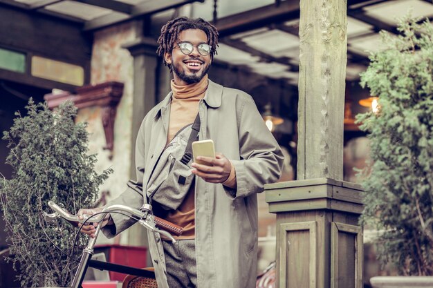 Mode de rue. Enthousiaste jeune homme gardant le sourire sur son visage en se tenant debout près de vélo