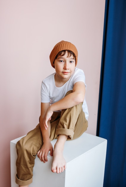 À la mode séduisant garçon tween aux cheveux noirs en chapeau assis sur un cube blanc en studio, la modélisation des enfants