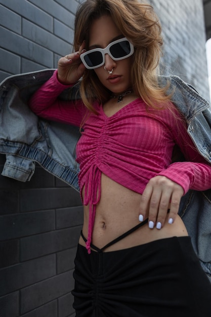 Mode sexy belle femme rousse hipster en streetwear élégant avec une veste en jean haut rose et une jupe noire se dresse et porte des lunettes de soleil blanches fraîches près d'un mur de briques noires dans la ville