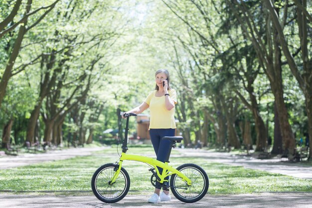 Mode de vie actif Une belle femme âgée se promène dans le parc monte un vélo jaune parle au téléphone Debout avec un vélo dans ses mains souriant s'amusant