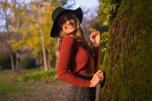 Mode de vie blonde fille de race blanche dans un pull rouge et un chapeau noir profiter de la nature dans un parc avec des arbres dans le beau portrait d'automne de la jeune femme à côté d'un arbre avec de la mousse