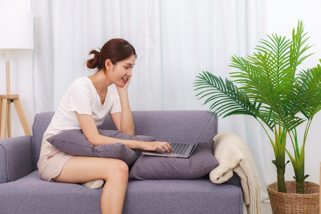 Mode de vie dans le concept de salon Jeune femme asiatique assise avec la main sur le menton et utilisant un ordinateur portable