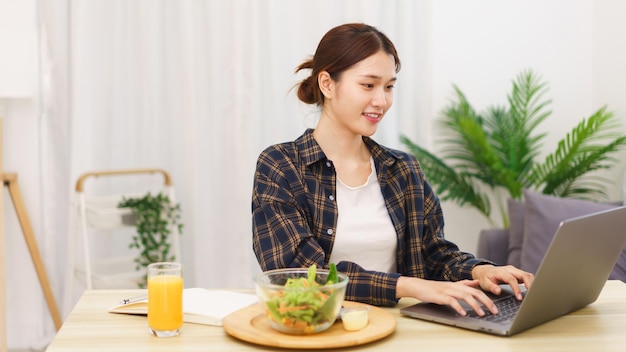 Mode de vie dans le concept de salon Jeune femme asiatique mangeant une salade de légumes et travaillant sur un ordinateur portable