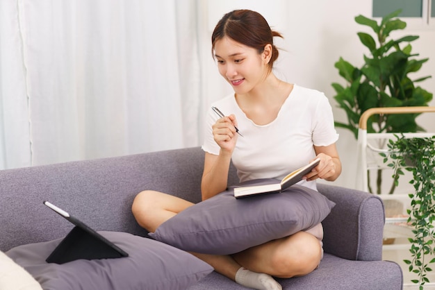 Mode de vie dans le concept de salon Jeune femme asiatique regardant sur une tablette et écrivant des données sur un ordinateur portable