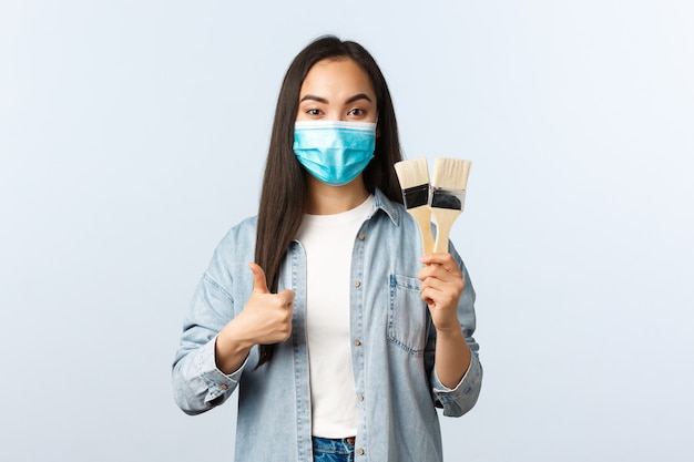 Mode de vie à distance sociale, pandémie de covid-19, concept de loisirs d'auto-isolement. Jolie femme asiatique portant un masque médical ayant l'air satisfaite, achetant des pinceaux en magasin pour rénovation, pouce levé