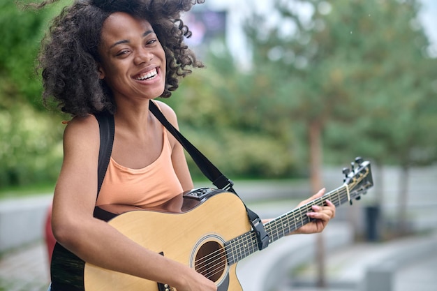 Mode de vie. Heureuse femme aux cheveux bouclés à la peau sombre avec un sourire à pleines dents avec une guitare regardant la caméra à l'extérieur par une belle journée chaude