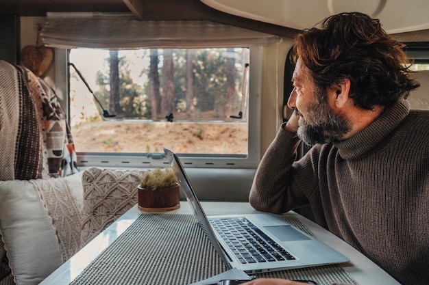 Mode de vie hors réseau avec un homme mûr travaillant sur un ordinateur portable à l'intérieur d'un camping-car et regardant la forêt à l'extérieur de la fenêtre Concept de personnes de la vie de van Travailleur à distance utilisant une connexion en ligne pour ordinateur