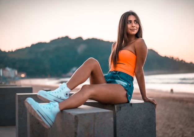 Mode de vie, une jeune brune assise dans un haut orange en regardant le coucher du soleil sur la plage