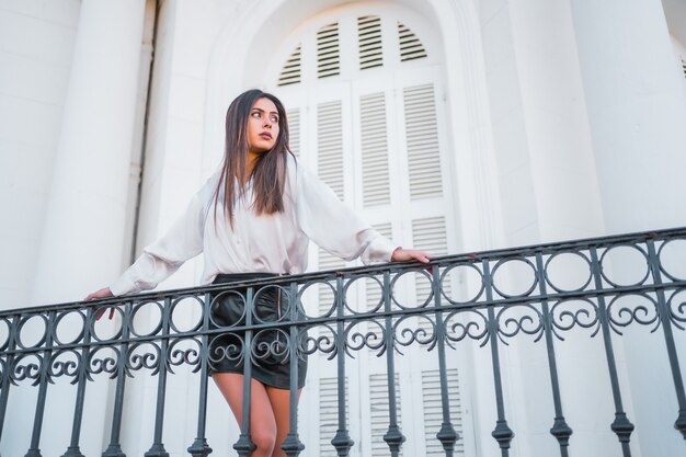 Mode de vie, une jeune brune vêtue d'une jupe en cuir noir et d'un T-shirt blanc, regardant le paysage depuis le balcon