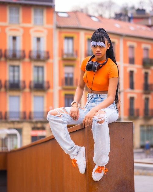 Mode de vie avec une jeune danseuse de trap avec des tresses. Fille noire d'ethnie africaine avec un t-shirt orange et un pantalon de cowboy. Avec des maisons