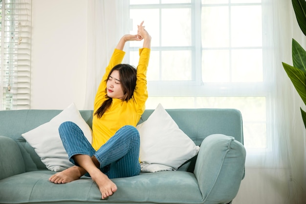 Mode de vie Jeune femme asiatique étirant ses bras sur un canapé dans le salon à la maison, activité de détente féminine heureuse se sentant paresseuse le week-end sur un canapé, pause sieste au repos