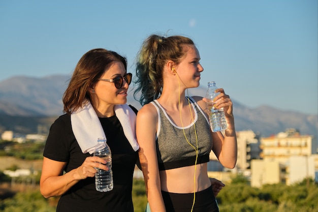 Mode de vie sain et actif, deux femmes adolescentes mère et fille en vêtements de sport parlant et buvant de l'eau après l'entraînement, fond de montagne, coucher de soleil
