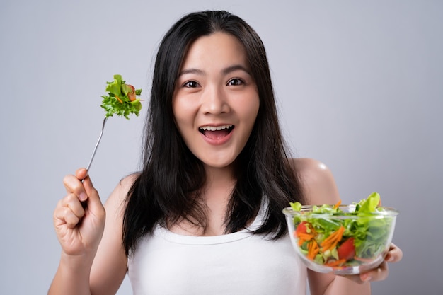 Mode de vie sain avec le concept de nourriture propre. Heureuse femme asiatique mangeant une salade isolée sur un mur blanc.