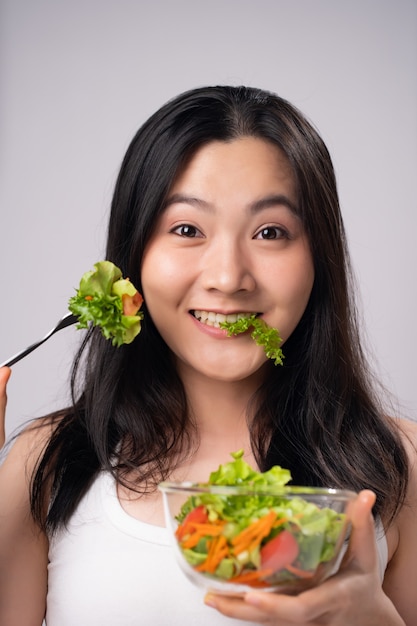 Mode de vie sain avec le concept de nourriture propre. Heureuse femme asiatique mangeant une salade isolée sur un mur blanc.