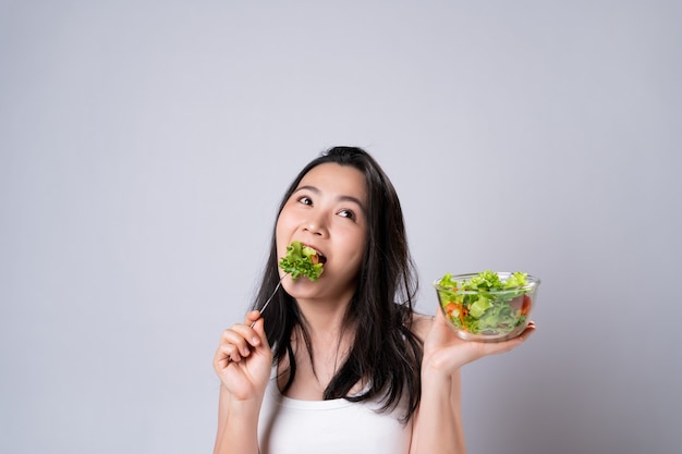 Mode de vie sain avec le concept de nourriture propre. Heureuse femme asiatique mangeant une salade isolée sur un mur blanc.