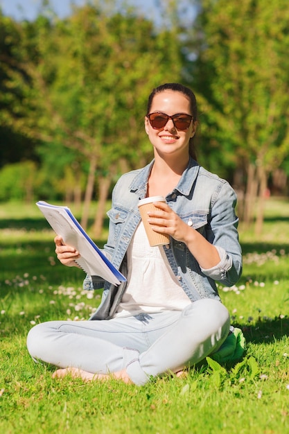 mode de vie, vacances d'été, éducation, boissons et concept de personnes - jeune fille souriante lisant des notes de cahier et buvant du café dans le parc