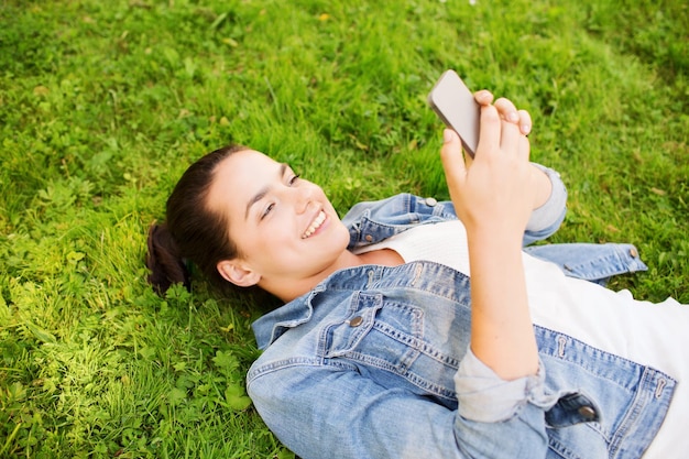 mode de vie, vacances d'été, technologie, loisirs et concept de personnes - jeune fille souriante avec un smartphone allongé sur l'herbe dans le parc