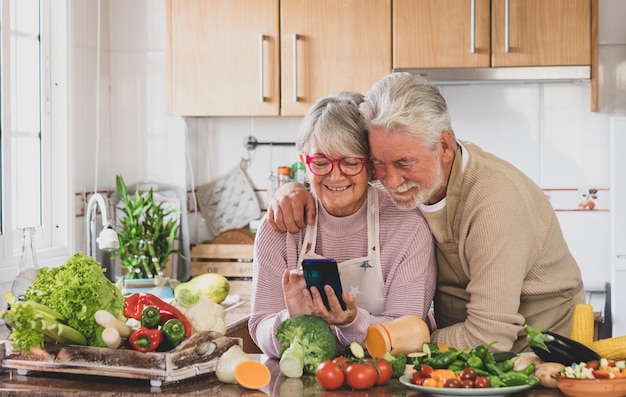 Mode de vie végétarien. Un beau couple de retraités aux cheveux blancs dans la cuisine regarde ensemble le smartphone. Sur la table un mélange de crudités de saison