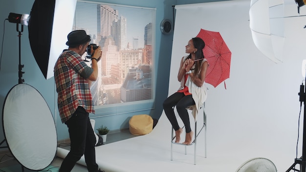 Modèle africain élégant posant avec un parapluie rouge sur une chaise haute de bar pour une séance photo de magazine de mode