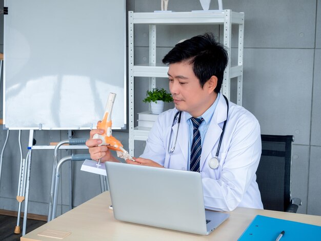 Modèle d'anatomie du squelette du pied de l'articulation de la cheville en gros plan tenant par un médecin asiatique orthopédique masculin avec une vidéoconférence d'ordinateur portable avec un patient dans un cabinet médical Télémédecine avec concept de médecin