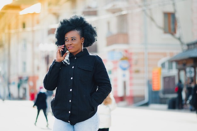 Modèle attrayant à la peau foncée heureuse avec une coiffure afro et un anneau dans le nez, posant à l'extérieur