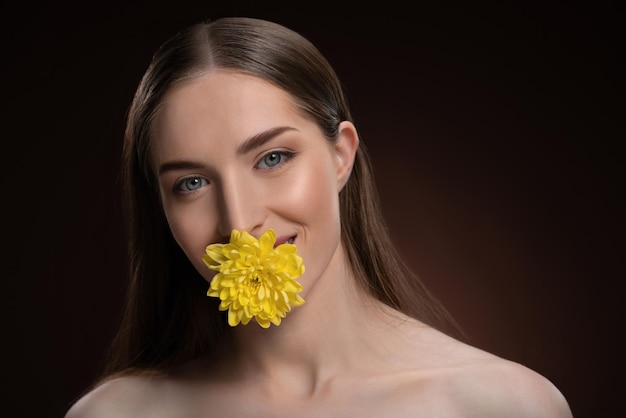 Modèle de beauté souriante aux épaules nues tenant une fleur dans sa bouche avec une peau de beauté et un long h...