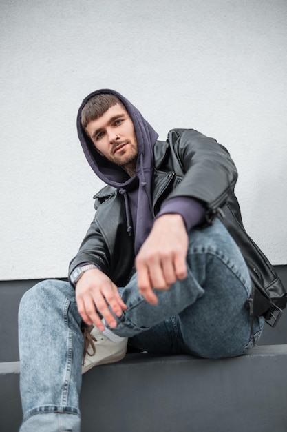 Modèle de bel homme à la mode cool avec une veste en cuir à capuche et un jean à la mode rock se trouve près d'un mur blanc dans la rue