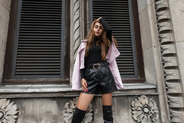 Modèle de belle femme à la mode dans des vêtements noirs élégants à la mode avec un short en cuir et des bottes hautes avec une veste rose posant près d'un bâtiment vintage dans une ville européenne