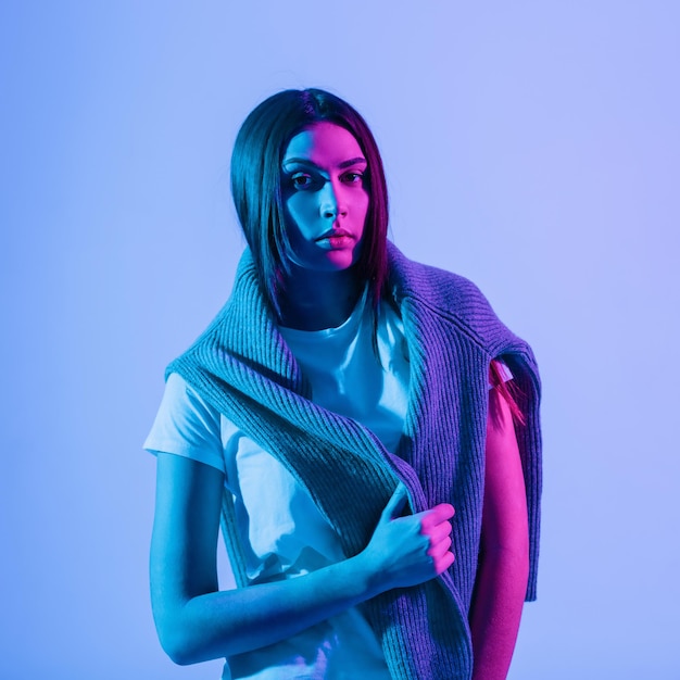 Modèle de belle fille aux cheveux bruns dans des vêtements décontractés à la mode avec un pull en tricot et un T-shirt blanc dans le studio avec des néons colorés et des lumières roses. Portrait féminin créatif coloré