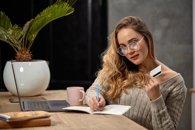 Modèle blogueur en pull beige à lunettes souriant assis dans la cuisine à table parlant par téléphone tenant ...