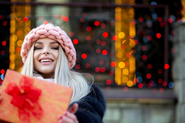 Un modèle blond expressif portant un manteau de fourrure et un bonnet tricoté vous tire une boîte-cadeau. Espace pour le texte