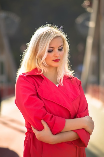 Modèle blonde attrayante avec le maquillage lumineux posant dans le manteau rouge à la rue