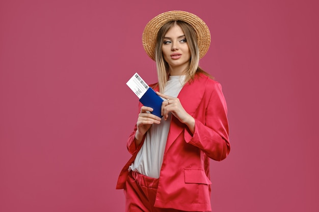 Modèle blonde en blouse blanche avec chapeau de paille et tailleur-pantalon rouge, elle tient un passeport et un billet