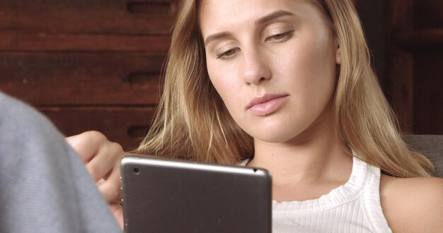 Modèle blonde chaude lisant sur sa tablette assise sur un canapé gris foncé recouvert d'un plaid doux